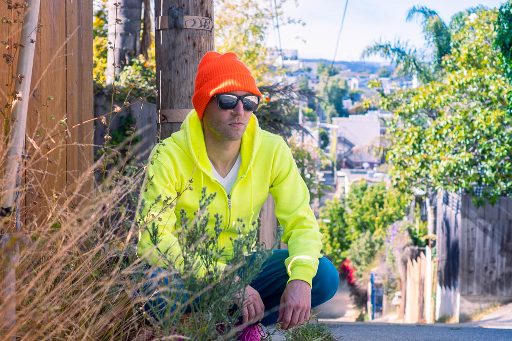 portrait of Marc wearing neon, kneeling in a sunlit alley